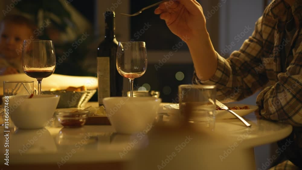 A warm family dinner setting features a hand serving food from a bowl, with a glass of wine and a baby playing in the background under soft evening lighting A warm family dinner setting features a hand serving food from a bowl, with a glass of wine and a baby playing in the background under soft evening lighting