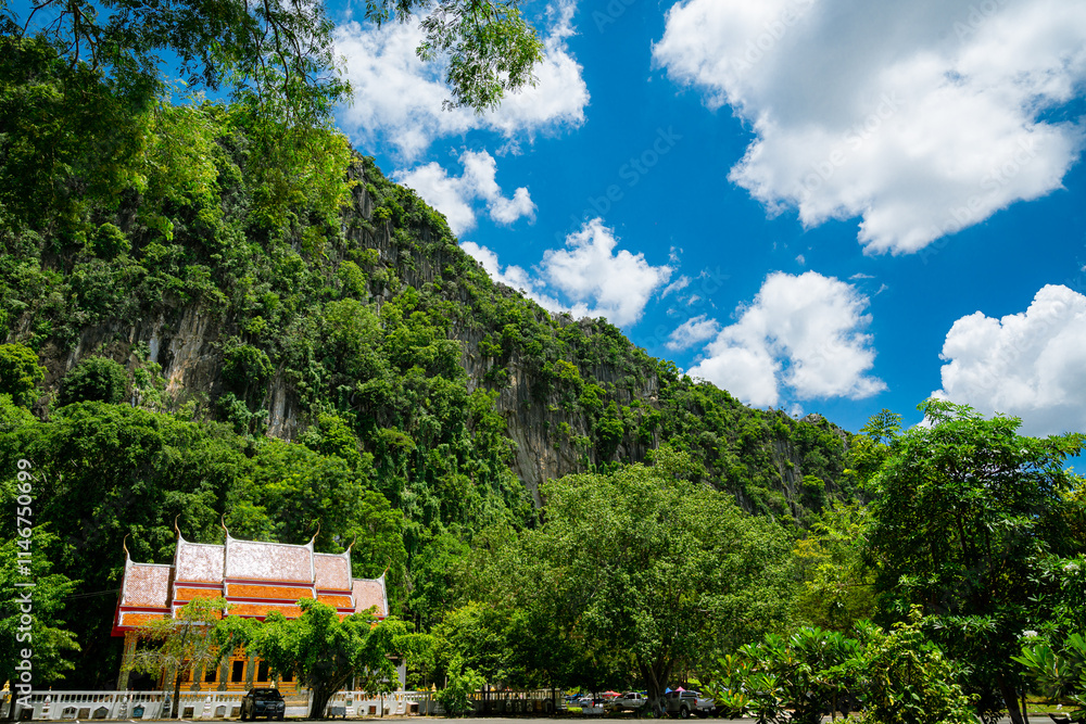 Fototapeta premium A rocky cliffs covered in lush green vegetation with orange roof of Wat Tum Khao Chakan, Sa Kaeo, Thailand