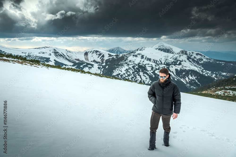 Stylish young man with a beard hiking in snowy mountain area wearing cold weather accessories, warm down jacket against a scenic backdrop of snowy mountains and snow-laden clouds. Stylish young man with a beard hiking in snowy mountain area wearing cold weather accessories, warm down jacket against a scenic backdrop of snowy mountains and snow-laden clouds.