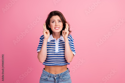 Young woman with cross fingers, standing against pink background in trendy striped top. Expressing hope and anticipation