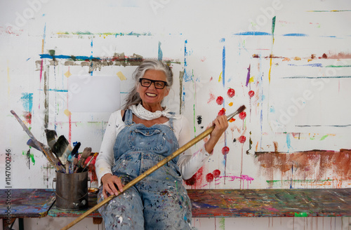 Crazy older mature woman artist, gray hair and glasses sits joyfully at a colorful painted wall, holding a large brush. Her denim overalls reflect a life filled with creativity and passion