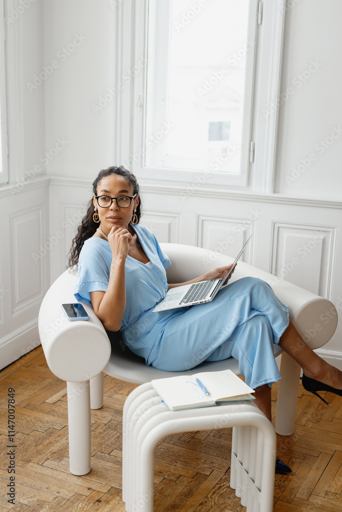 African-American woman working on laptop while seated in modern chair in bright office environment during daylight. African-American woman working on laptop while seated in modern chair in bright office environment during daylight.