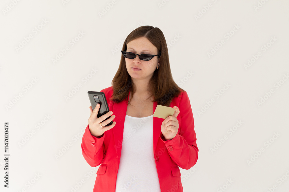 Cheerful young girl in black sunglasses showing plastic credit card while using mobile phone isolated over white background, wearing red jacket Cheerful young girl in black sunglasses showing plastic credit card while using mobile phone isolated over white background, wearing red jacket