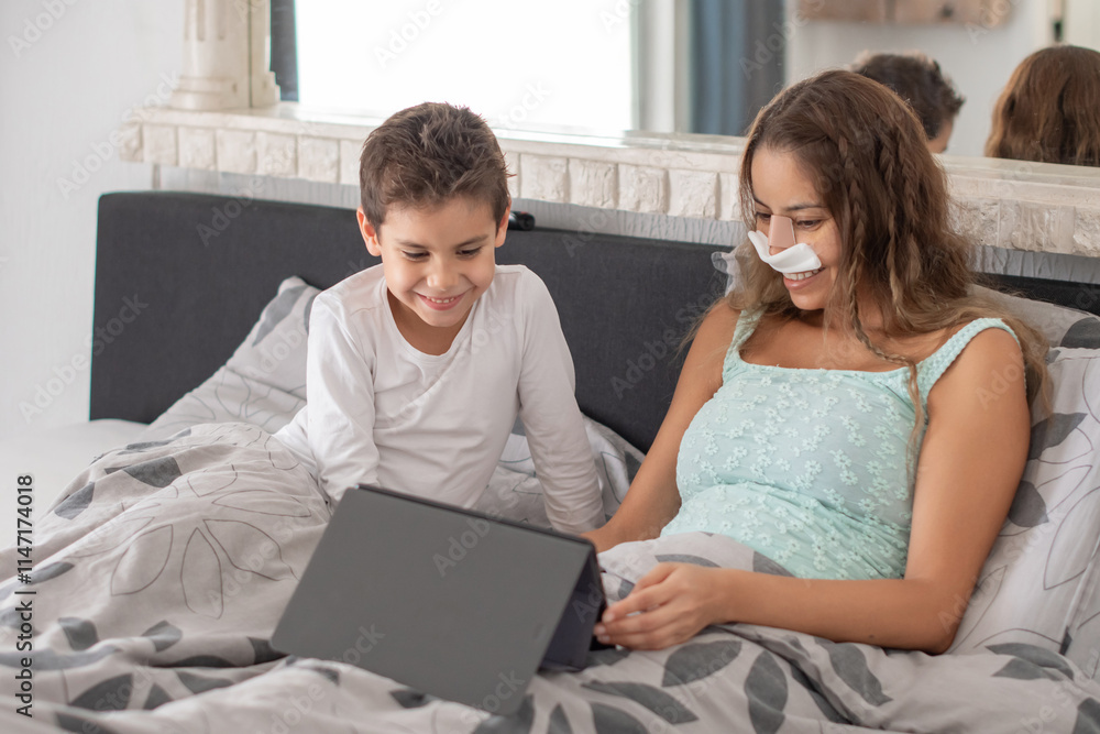 Smiling mother with bandage on nose lying in cozy bed and helping her schoolboy son to do homework, concept of upbringing and education, family relaxing lying among pillows, Smiling mother with bandage on nose lying in cozy bed and helping her schoolboy son to do homework, concept of upbringing and education, family relaxing lying among pillows,