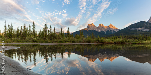 Three Sisters in Canmore seen at golden hour, sunset on blue sky day, afternoon with calm, peaceful reflection in water below famous, tourist, tourism mountains, area summer.	