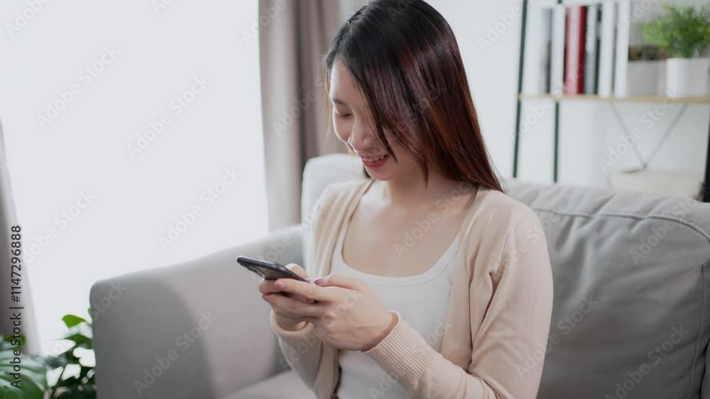 Beautiful and lovely Asian woman is using a smartphone with a happy feeling, sitting on a sofa in a living room at home. Using technology at home concept. Beautiful and lovely Asian woman is using a smartphone with a happy feeling, sitting on a sofa in a living room at home. Using technology at home concept.