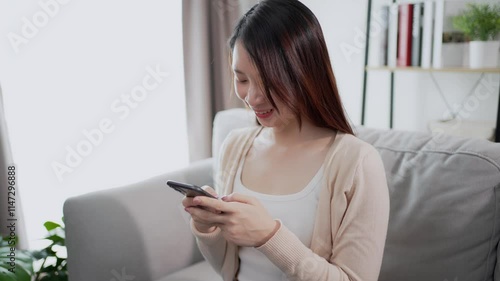 Beautiful and lovely Asian woman is using a smartphone with a happy feeling, sitting on a sofa in a living room at home. Using technology at home concept.