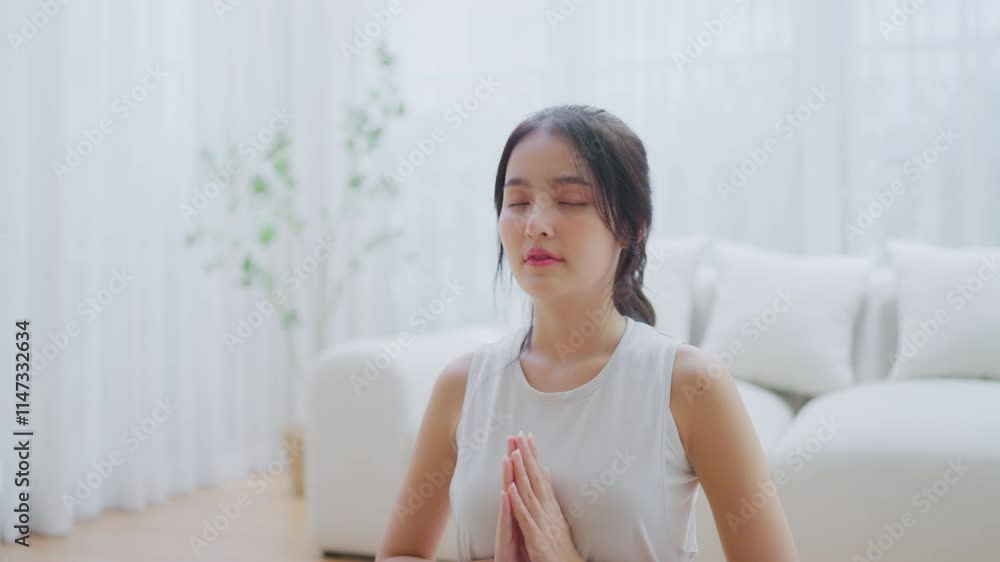 Happy young Asian woman practicing yoga and meditation at home sitting on floor in living room in lotus position and relaxing with closed eyes. Mindful meditation and wellbeing concept Happy young Asian woman practicing yoga and meditation at home sitting on floor in living room in lotus position and relaxing with closed eyes. Mindful meditation and wellbeing concept