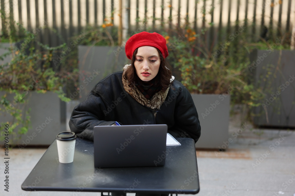 A young woman in a red beret works on her laptop at an outdoor caf, enjoying her coffee A young woman in a red beret works on her laptop at an outdoor caf, enjoying her coffee