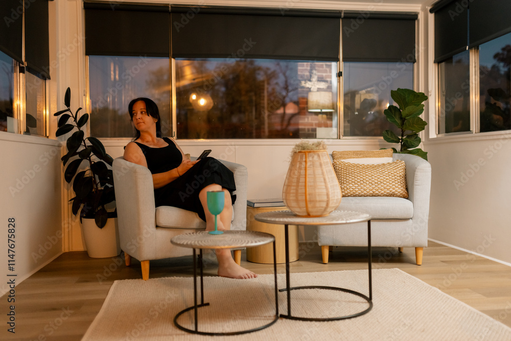© Austockphoto - Woman in black dress looking out window from cosy armchair in warmly lit room