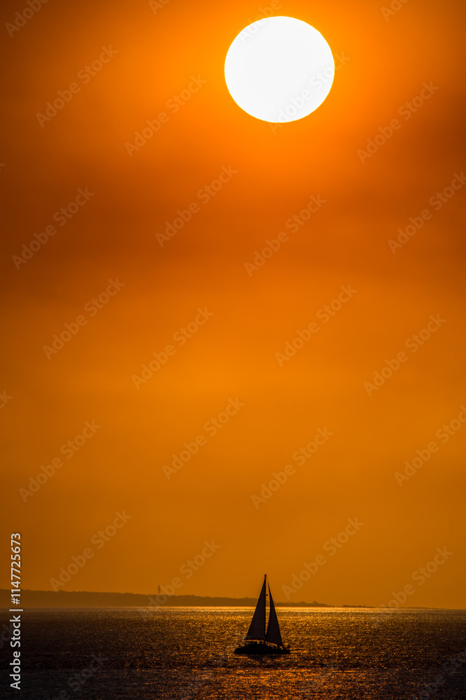 © Austockphoto - A bright sun hangs low in the sky with a small silhouette of a sailboat on the ocean. © Austockphoto - A bright sun hangs low in the sky with a small silhouette of a sailboat on the ocean.