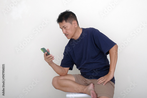 Asian guy in casual clothes, sitting alone on white stool reading messages on his smartphone photo