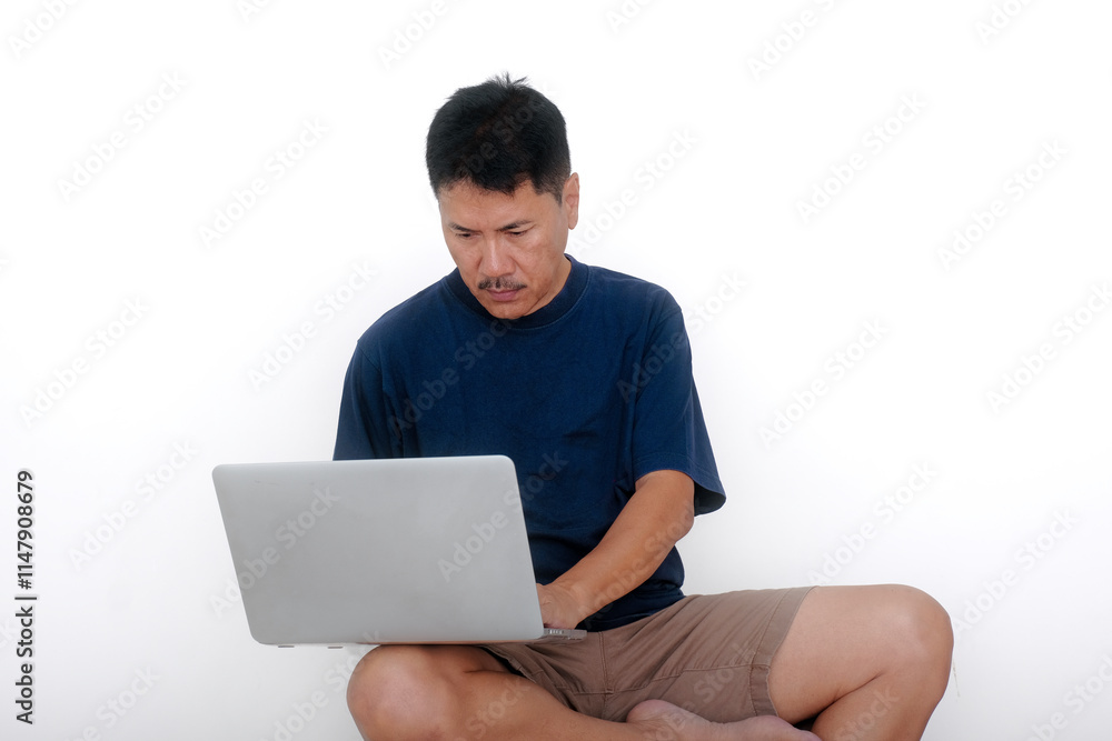 Asian guy in casual clothes, sitting alone on white stool working on his laptop computer Asian guy in casual clothes, sitting alone on white stool working on his laptop computer