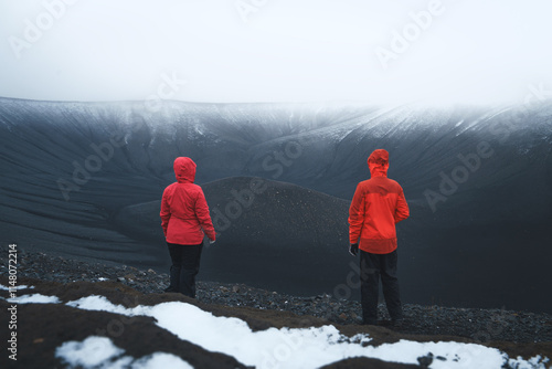 A hiking couple in red jackets standing at the edge of the Hverfjall volcano, admiring the black tuff ring in snowy and misty weather photo