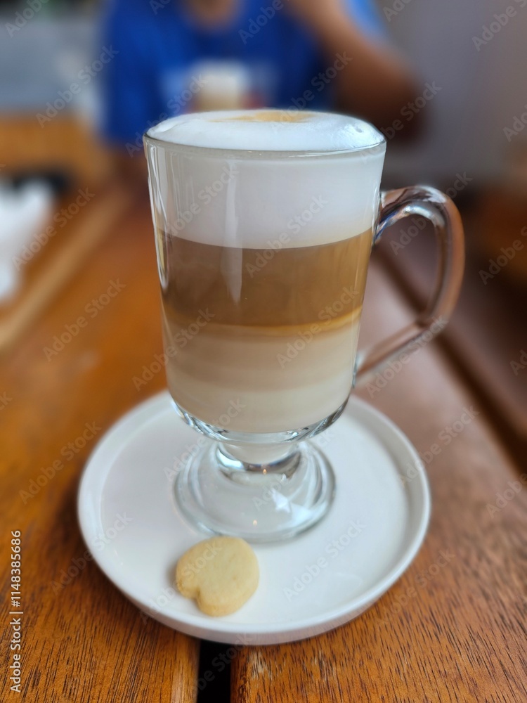 Layered coffee latte in a glass mug with a lot of foam and a small cookie with the shape of a heart Layered coffee latte in a glass mug with a lot of foam and a small cookie with the shape of a heart