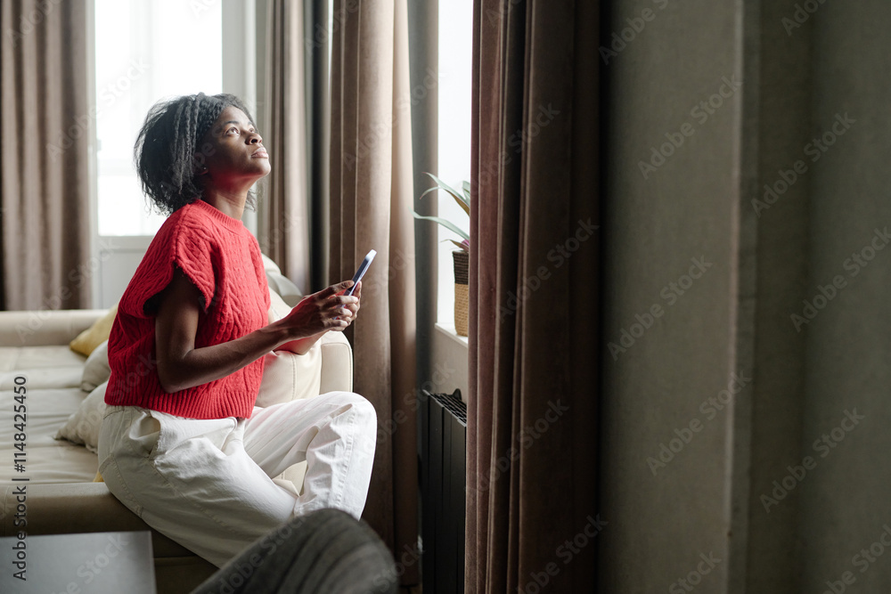 African American woman sitting on couch near huge window while using smartphone in her free time