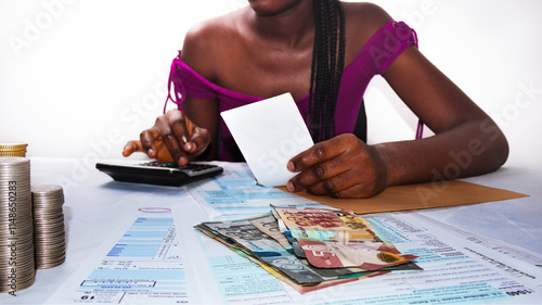 Young black african female lady woman wearing Purple top sitting at office desk holding paper recipt pressing calculator calculating with Ghanaian cedi notes in front on table photo