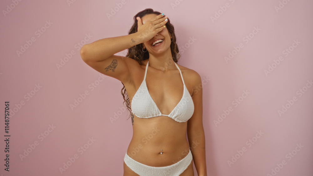 Beautiful young hispanic woman in a white bikini posing with a playful smile, covering her eyes against a pink background wall. Beautiful young hispanic woman in a white bikini posing with a playful smile, covering her eyes against a pink background wall.