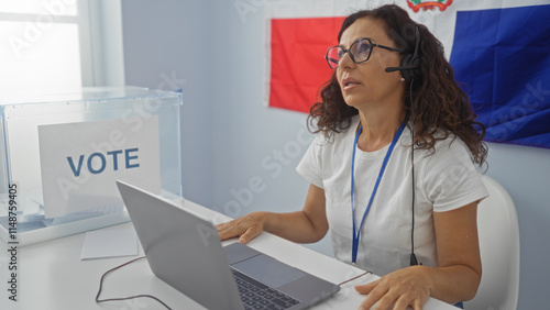 Fotografie Woman working at an electoral office in the dominican republic, wearing a headset and sitting by a voting box and laptop with a dominican flag in the background