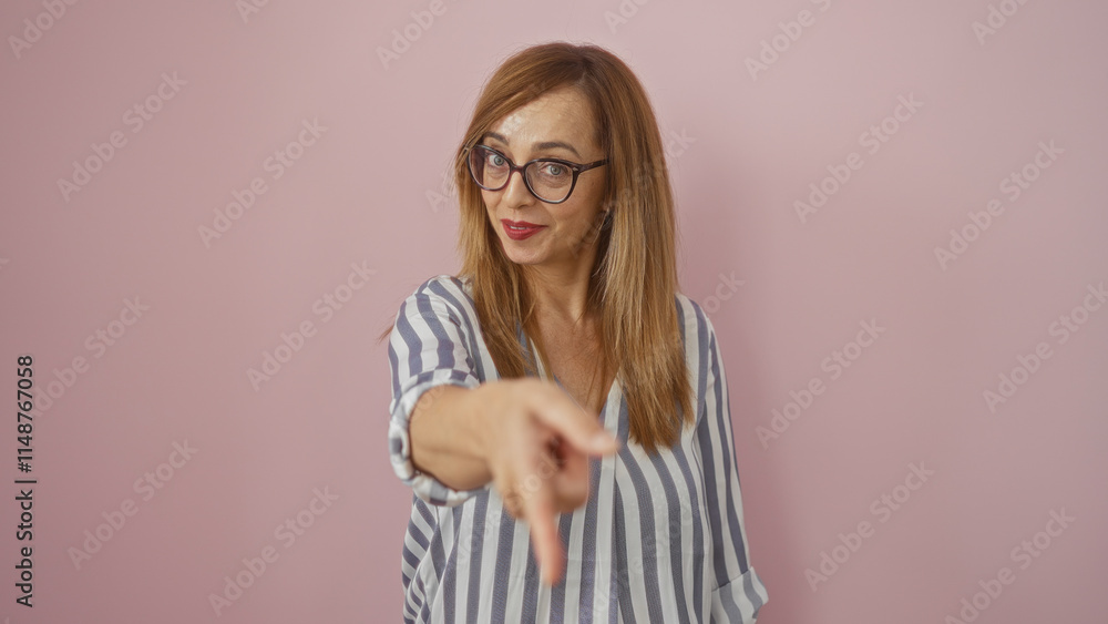 Blonde woman pointing forward confidently against a pink wall wearing glasses and a striped shirt Blonde woman pointing forward confidently against a pink wall wearing glasses and a striped shirt