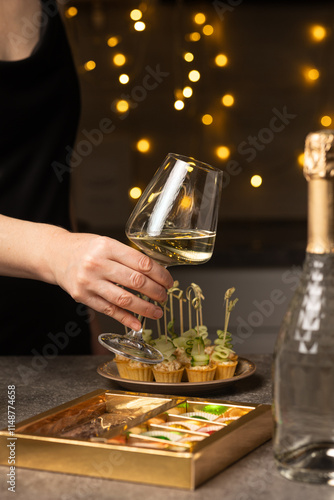 A woman holds a glass of wine in her hand in the kitchen. There is a bottle of champagne and a snack on the table. Vertical photo. photo