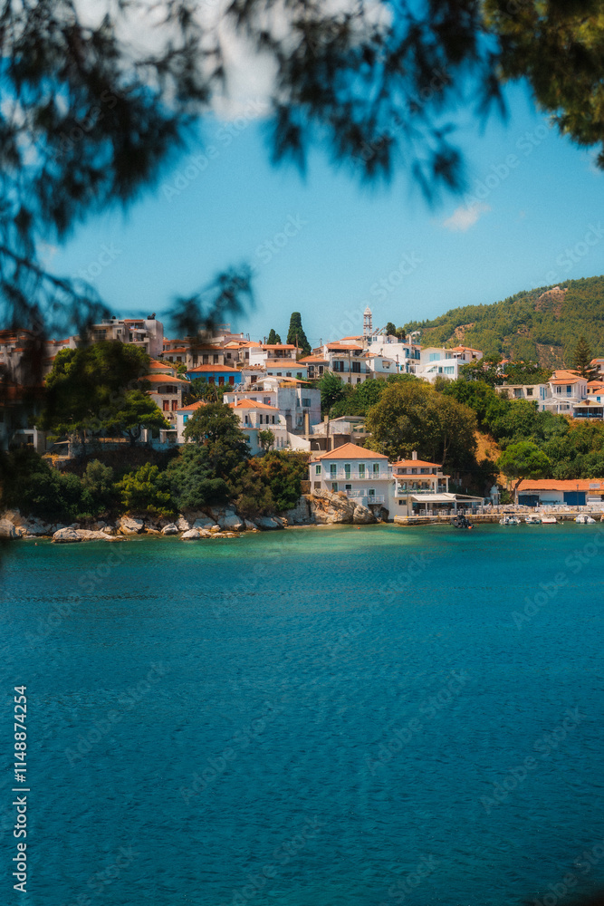 Obraz premium Whitewashed houses cluster on a hillside, overlooking a beautiful bay with crystal-clear turquoise water, framed by lush greenery under a vibrant blue sky