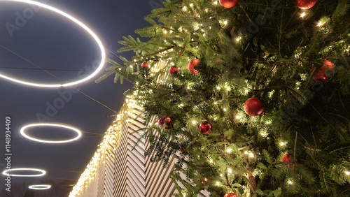 Closeup of festive fir tree branch decorated with red glass balls and lights garland on outdoor Christmas market at holiday evening or night. With no people traditional Xmas illumination concept.