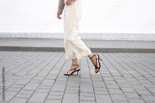 Woman in white denim pants and black heeled sandals walking outside along a city street covered with paving slabs. Trendy elegant casual outfit. Street fashion. Women's legs, without a face.