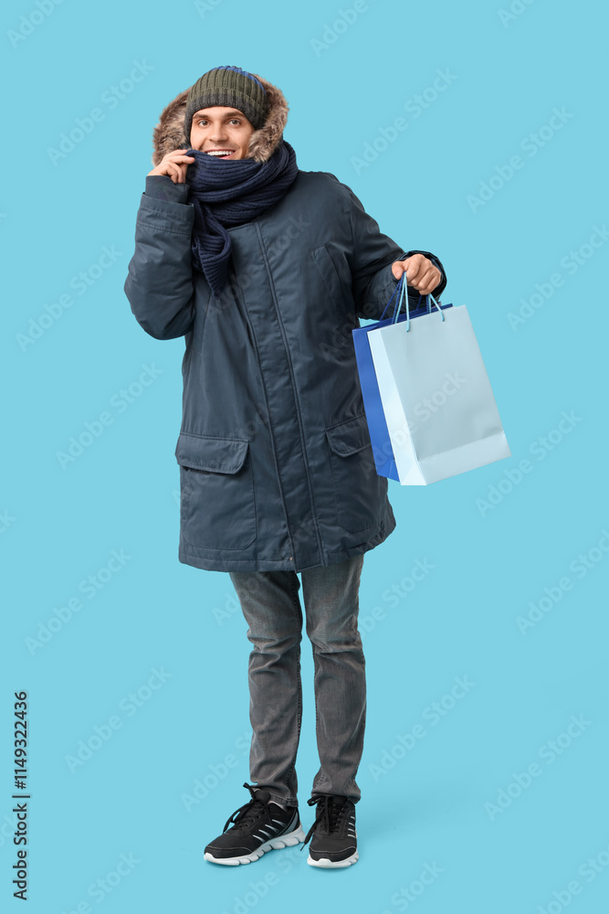 Young man in stylish puffer jacket and scarf with shopping bags on blue background Young man in stylish puffer jacket and scarf with shopping bags on blue background
