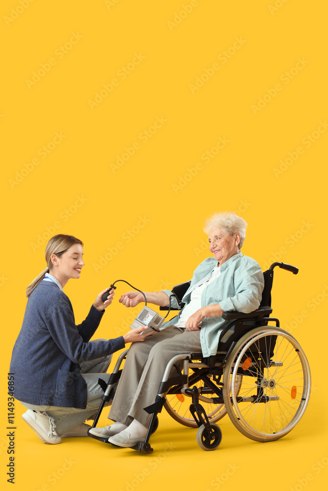 Senior woman in wheelchair with nurse measuring blood pressure on yellow background Senior woman in wheelchair with nurse measuring blood pressure on yellow background