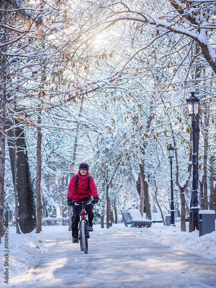 A man rides his bicycle in a winter park. A guy in a helmet and red jacket on a red bicycle riding on a sunny winter day A man rides his bicycle in a winter park. A guy in a helmet and red jacket on a red bicycle riding on a sunny winter day