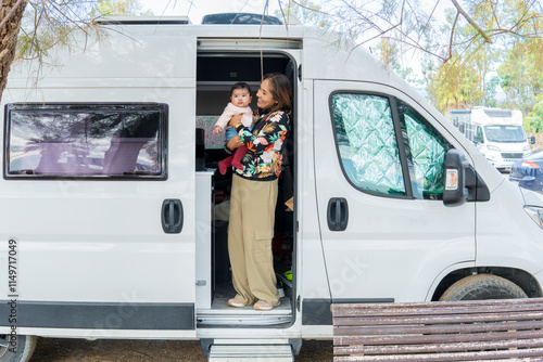 beautiful mother and her baby in her modern camper van come out of the sliding door at a campsite. Concept: camper van family vacation, camper van family tourism, camper life with babies.