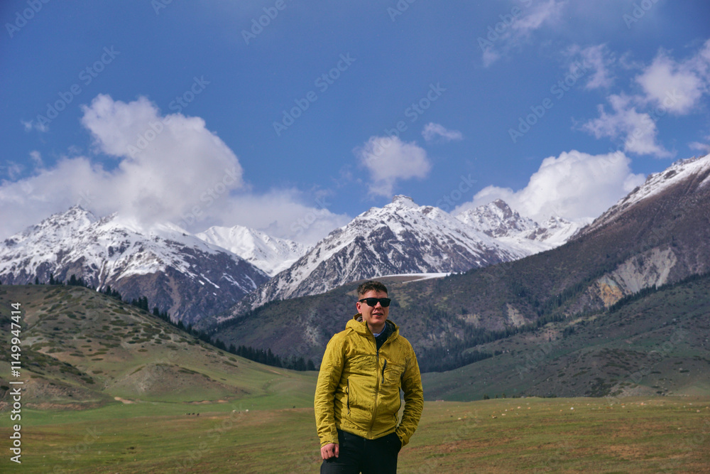 A young man in sunglasses stands against the backdrop of snow-capped mountain peaks. Trekking in the mountains A young man in sunglasses stands against the backdrop of snow-capped mountain peaks. Trekking in the mountains