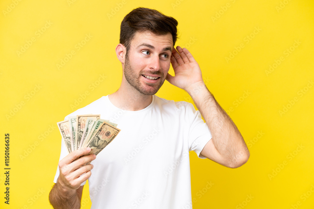 Young caucasian man taking a lot of money isolated on yellow background listening to something by putting hand on the ear Young caucasian man taking a lot of money isolated on yellow background listening to something by putting hand on the ear