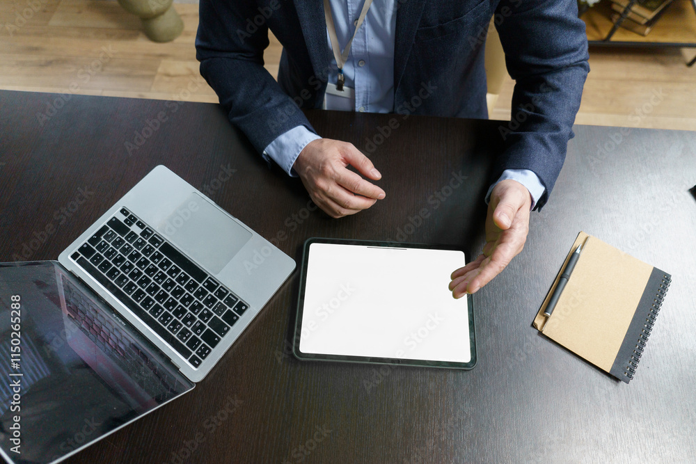 Fototapeta premium Man is sitting at a desk with a laptop and a tablet. The tablet is open to a blank screen