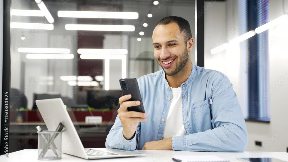 Satisfied happy african american businessman received great news on phone while sitting at workplace in business office. Smiling glad black man worker reads a pleasant message on smartphone, rejoices Satisfied happy african american businessman received great news on phone while sitting at workplace in business office. Smiling glad black man worker reads a pleasant message on smartphone, rejoices