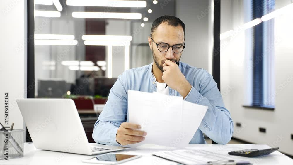 Confused african american businessman having difficulty with paper work sitting at desk at workplace in business office. Puzzled entrepreneur looks at the documents and cannot understand the problem Confused african american businessman having difficulty with paper work sitting at desk at workplace in business office. Puzzled entrepreneur looks at the documents and cannot understand the problem