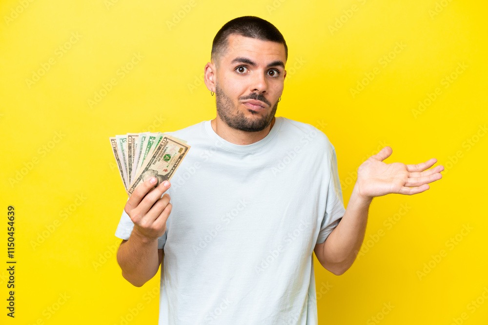 Young caucasian man taking a lot of money isolated on yellow background having doubts while raising hands Young caucasian man taking a lot of money isolated on yellow background having doubts while raising hands