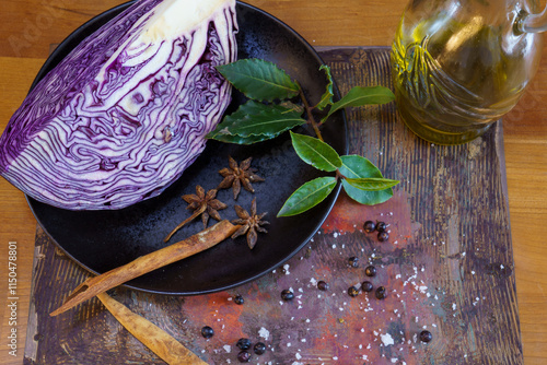 A quarter of a red cabbage head on a black plate with the spices star anise, bay leaf, cinnamon, juniper berries and salt crystals. Next to it a glass carafe with olive oil photo