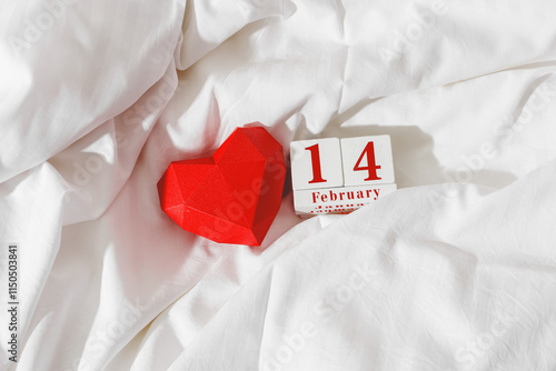 фотографія Valentine's Day concept, red paper valentine heart and 14 february holiday date on wooden calendar in bed on on  white crumpled sheets