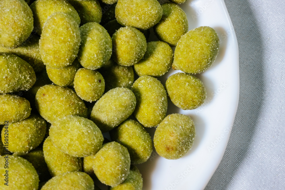 A close-up of a white plate filled with bright yellow-green wasabi-coated nuts. The nuts are coated in a fine powder, giving them a crunchy and spicy appeal. A close-up of a white plate filled with bright yellow-green wasabi-coated nuts. The nuts are coated in a fine powder, giving them a crunchy and spicy appeal.