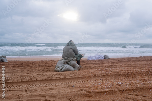 A tired man is sitting on a sack by the sea, dressed in a protective suit. Puts fuel oil in a bag. An environmental disaster. Elimination of oil spill. photo