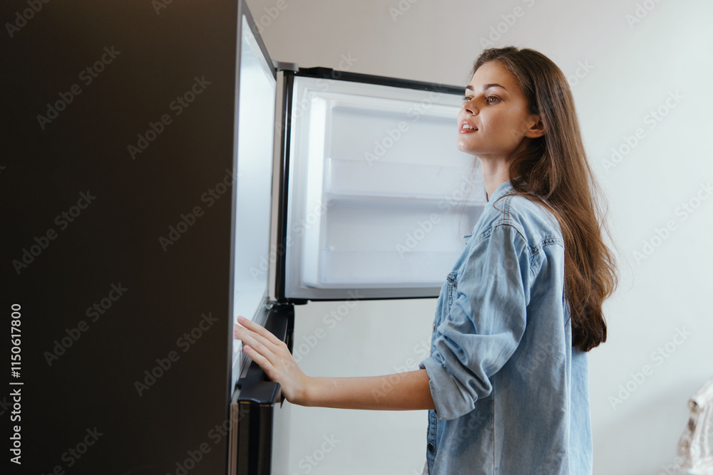 Young woman in denim shirt looking into an open refrigerator, expressing curiosity and excitement about healthy food options Young woman in denim shirt looking into an open refrigerator, expressing curiosity and excitement about healthy food options