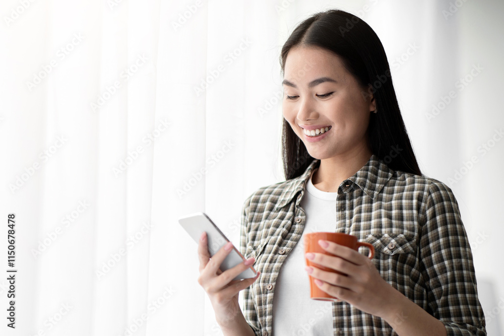 Beautiful asian young woman with coffee mug using mobile phone, standing next to window, panorama with copy space. Cheerful lady drinking tea, chatting with boyfriend on smartphone and smiling