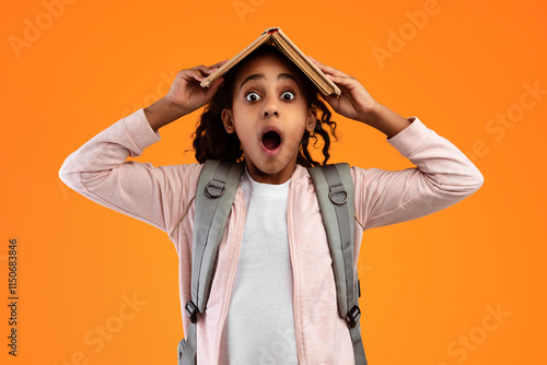 Reading Programs. Portrait of funny African American student girl putting and holding academic book on her head and looking at camera, wearing backpack, having fun, yellow color studio background photo