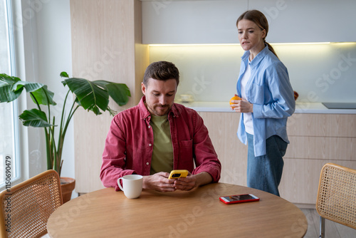 Fotografie Woman secretly reads husband messages over shoulder while he chats on kitchen