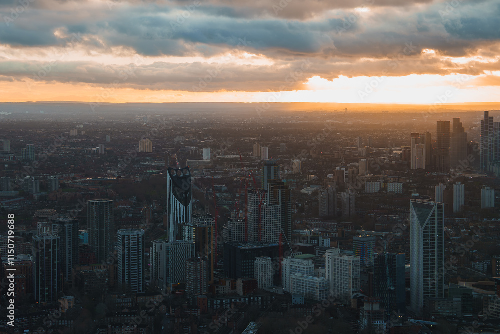 Fototapeta premium Aerial view of London during sunset, showcasing modern skyscrapers like Strata SE1. The cityscape is illuminated by a warm glow against dark clouds.