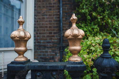 Close up of gold painted finials on a black iron fence with a brick building and green foliage, typical of London's historic decorative ironwork. photo