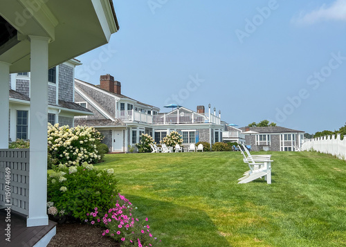 A sunny lawn with white Adirondack chairs and blooming hydrangeas leading to a beach house with a fence in the background, perfect for relaxing and enjoying a coastal view.
