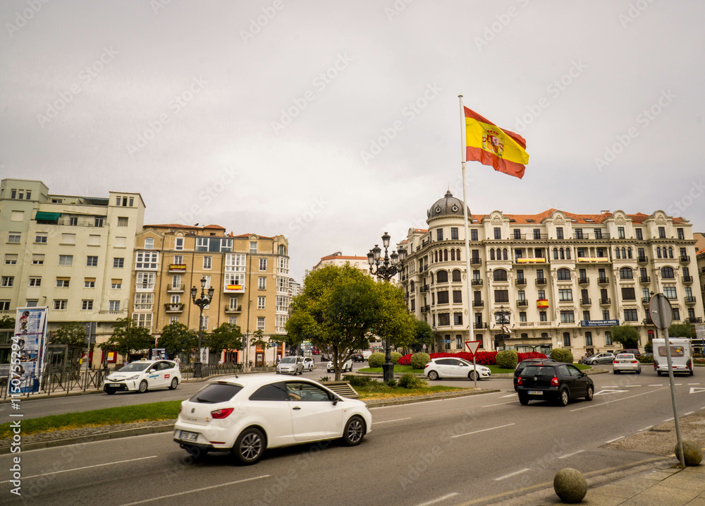 Fototapeta premium Large Spanish Flag Waving on an Avenue in the City of Santander with Cars Driving and Buildings in the Background with Flags Hanging on Balconies